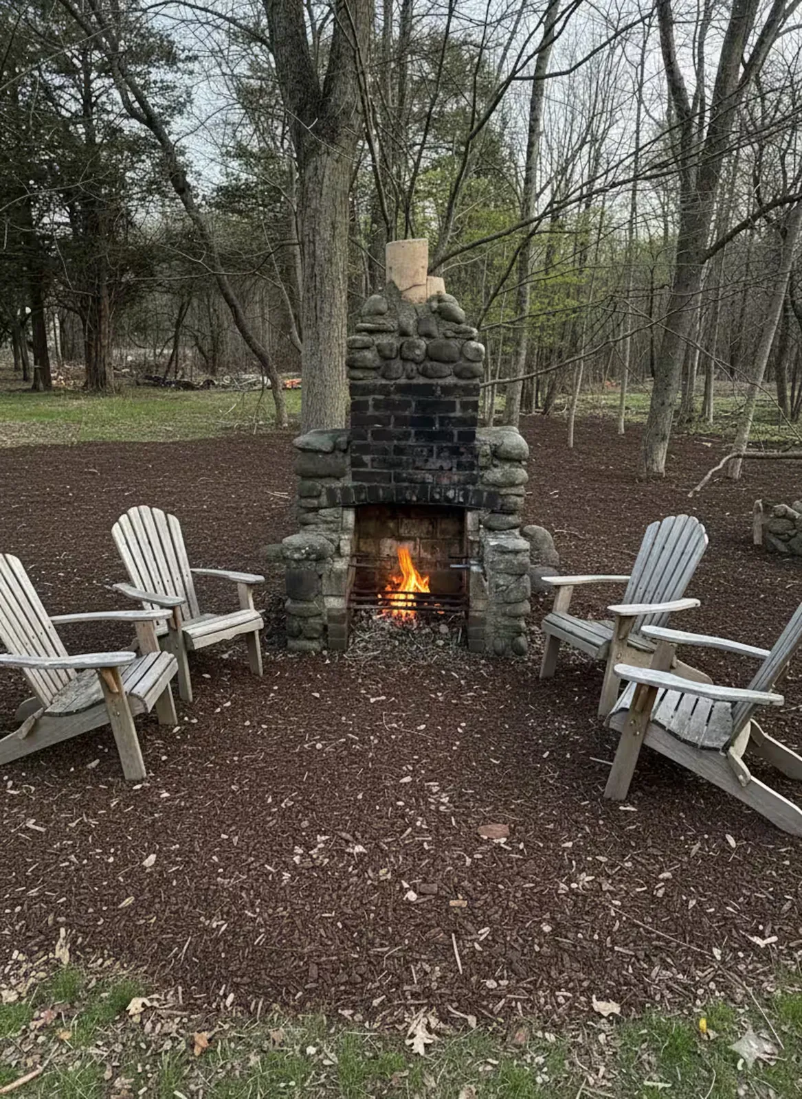 Outdoor stone fireplace lit at dusk, ringed by Adirondack chairs